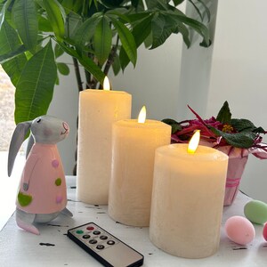 Könnte beinhalten: Drei weiße Kerzen in Säulenform mit flackernden Flammen, eine rosafarbene Hasenfigur und eine Fernbedienung auf einem weißen Tisch mit dekorativem Design.