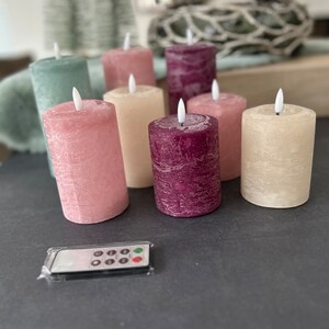 May include: Set of eight battery-operated pillar candles with a flickering flame effect. The candles are in various colors, including pink, cream, green, and burgundy. The candles are arranged on a dark surface with a remote control.