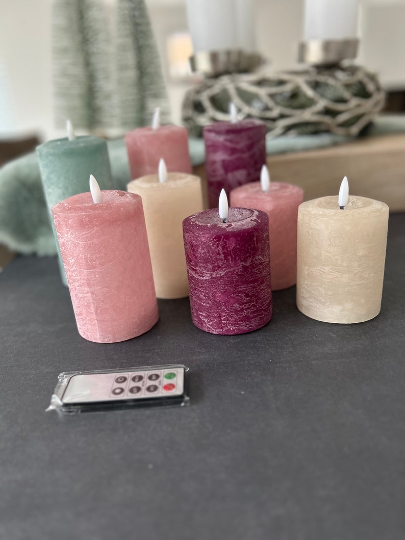 May include: Set of eight battery-operated pillar candles with a flickering flame effect. The candles are in various colors, including pink, cream, green, and burgundy. The candles are arranged on a dark surface with a remote control.
