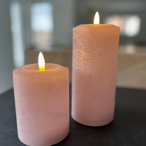 May include: Two pink pillar candles with a flickering flame on a black surface. The candles have a textured, frosted finish.