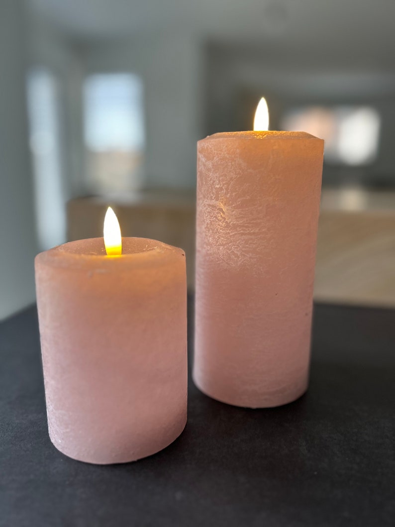 May include: Two pink pillar candles with a flickering flame on a black surface. The candles have a textured, frosted finish.