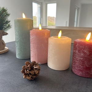 Könnte beinhalten: Vier Stumpenkerzen mit flackernden Flammen. Die Kerzen sind in den Farben Grün, Rosa, Creme und Burgunderrot. Die Kerzen stehen auf einer dunklen Oberfläche.