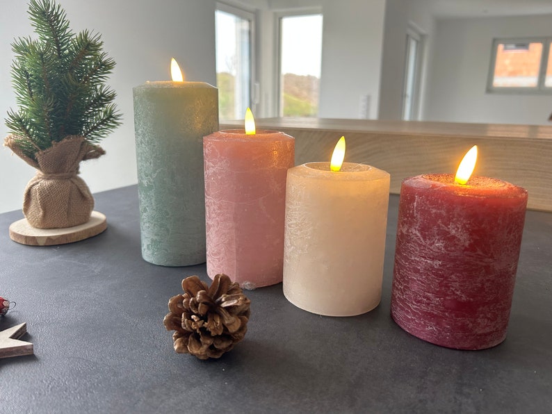 May include: Four pillar candles with flickering flames. The candles are in shades of green, pink, cream, and burgundy. The candles are on a dark surface.