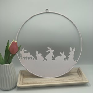 Könnte beinhalten: Weiße Ostergirlande aus Metall mit einem runden Design, das die Silhouette von fünf Hasen zeigt. Die Girlande hat oben eine Aufhängeschlaufe.