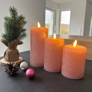 May include: Three pink pillar candles with a flickering flame effect. The candles are arranged in a row on a black surface. There is a small pine tree and some ornaments in the foreground.