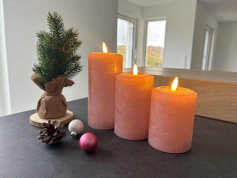 May include: Three pink pillar candles with a flickering flame effect. The candles are arranged in a row on a black surface. There is a small pine tree and some ornaments in the foreground.