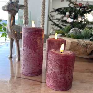 May include: Three red pillar candles with a textured finish and flickering LED flames. The candles are on a wooden surface.