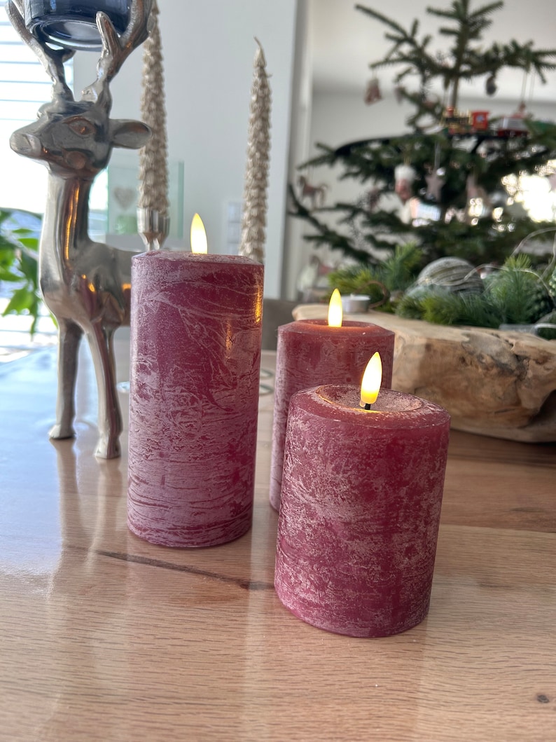 May include: Three red pillar candles with a textured finish and flickering LED flames. The candles are on a wooden surface.