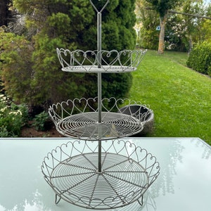 Könnte beinhalten: Ein dreistöckiger Kuchenständer aus Metall mit Herzform. Der Ständer ist aus Draht gefertigt und hat eine silberne Oberfläche.