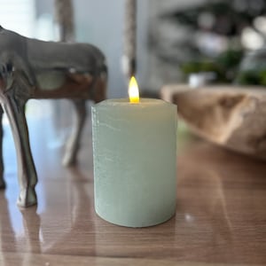 Könnte beinhalten: Eine tealfarbene Stumpenkerze mit flackernder Flamme. Die Kerze steht auf einer Holzoberfläche.