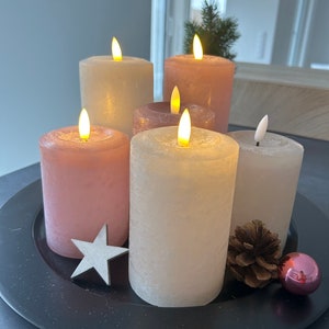 May include: Five pillar candles on a black tray. Two candles are white with a textured finish, and three candles are pink with a textured finish. The candles are lit and have a warm glow. There is a small pine cone and a pink ornament on the tray.