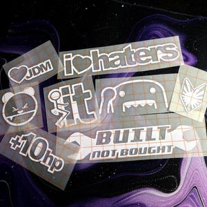 May include: A collection of white vinyl stickers with various designs and text. The stickers include phrases like "I ❤️ haters", "Built Not Bought", and "+10hp", along with other graphic elements. The stickers are on a purple and black background.