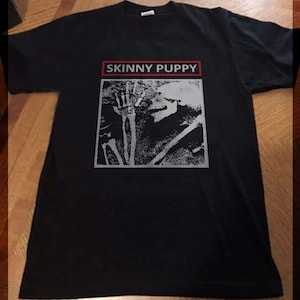 Puede incluir: Camiseta negra con un gráfico blanco de un esqueleto con el texto "SKINNY PUPPY" en rojo.