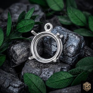 May include: A silver-toned metal pendant setting featuring a circular frame with four prongs designed to hold a gemstone. The setting includes a small circular loop at the top for attachment, showcasing a polished metallic finish and a structural design.