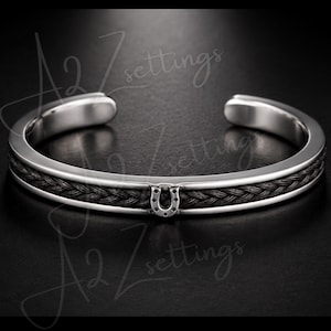 Può includere: Bracciale a polsino color argento con fascia centrale nera intrecciata e ciondolo a forma di ferro di cavallo. Il bracciale ha una finitura lucida e un design aperto. Lo sfondo ha il testo A2Z settings.