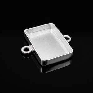 May include: A rectangular silver-coloured jewellery finding with a recessed interior. The finding has two small loops on opposite sides. The item is designed for jewellery making and crafting.