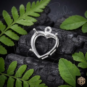 May include: A silver-colored heart-shaped pendant setting featuring a prong-style mount designed to hold a gemstone. The metal frame includes a small circular loop at the top for attachment to a chain or cord, showcasing a polished metallic finish.