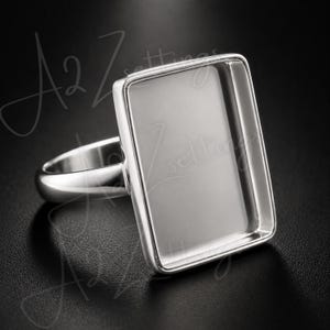 May include: A silver-toned metal ring featuring a large, empty rectangular bezel setting with raised edges. The ring has a smooth, polished band and a flat, open center designed for a stone or inlay, presented against a dark background.