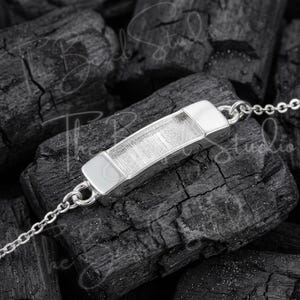 May include: A silver-toned bracelet featuring a central rectangular bar component with a textured, brushed metal finish. The bar is connected to a delicate chain on both sides, showcasing a polished, reflective frame around the center.