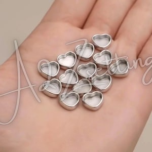 May include: A collection of small, heart-shaped metal settings. The settings are silver-colored with a smooth, reflective surface. The heart shapes are uniform in size and design, with a shallow, open interior. The settings are likely for jewelry making or crafting.