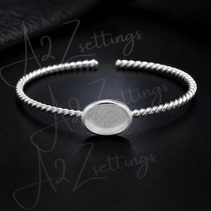 May include: A silver-toned cuff bracelet with a twisted rope design. The bracelet features an oval setting in the center, designed to hold a stone or other decorative element. The open design allows for easy adjustment and wear.