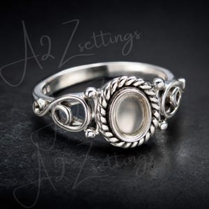 May include: A silver ring featuring an oval-shaped central setting surrounded by a twisted rope-style border. The band displays intricate scrollwork and filigree details on both sides of the center piece. The text A2 Zettings is visible.