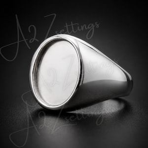 May include: A polished silver-tone signet ring featuring a smooth, oval-shaped flat bezel setting designed for customization. The ring has a solid, reflective metallic band and a clean, minimalist face. The text A2 Zettings is visible.