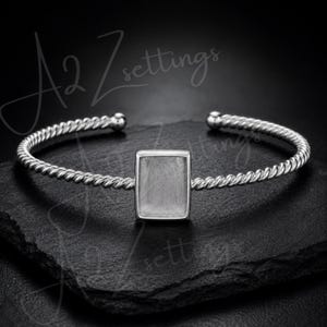 Può includere: Un bracciale a polsino argentato con incastonatura rettangolare. Il bracciale presenta un design a corda intrecciata e piccole estremità rotonde. Lo sfondo scuro esalta la lucentezza metallica. Il testo "A2 Zsettings" è visibile. Circonferenza di circa 16 cm.