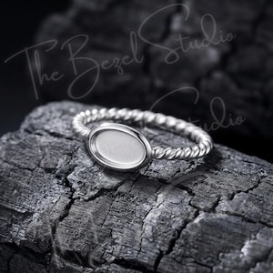 May include: A silver ring featuring a twisted rope-style band and a smooth, oval-shaped bezel setting. The ring has a polished metallic finish and includes the text The Bezel Studio overlaid on the image. The design is simple and circular.