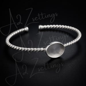 Op de afbeelding: Een zilverkleurige manchetarmband met een gedraaid touwontwerp. De armband heeft een ovale bezel-instelling in het midden, perfect voor het toevoegen van een edelsteen of ander decoratief element. De armband wordt tegen een donkere achtergrond getoond.