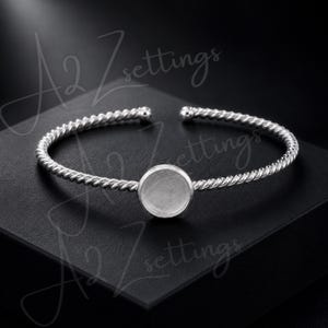May include: Silver-toned cuff bracelet with a twisted rope design. The bracelet features an open design and a circular setting in the center, perfect for adding a custom charm or stone. The bracelet is displayed on a black surface.