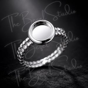 May include: A silver-toned ring featuring a circular, smooth-surfaced bezel setting designed for a stone or cabochon. The band consists of two parallel rows of small, polished metallic beads. The text The Bezel Studio is visible in the image.
