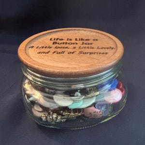 Peut inclure: Un bocal en verre transparent rempli de boutons colorés variés, surmonté d'un couvercle en bois rond. Le couvercle porte l'inscription gravée : Life is Like a Button Jar, A Little loose, a Little Lovely, and Full of Surprises. Le bocal est clair.