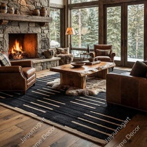 May include: A living room featuring a stone fireplace, a wooden coffee table, and brown leather furniture. A black and white patterned rug covers the wooden floor. A large chandelier hangs from the ceiling, and windows reveal a forest view.