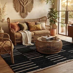 May include: A living room featuring a rattan sofa and armchair with tan cushions, a round rattan coffee table with a glass top, and a black area rug with white horizontal stripes. A large macrame wall hanging is displayed above the sofa.