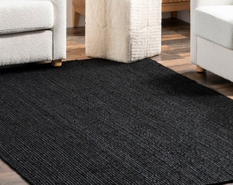 May include: A black woven rug with a textured surface. The rug is placed in a living room setting with two white armchairs and a white ottoman.