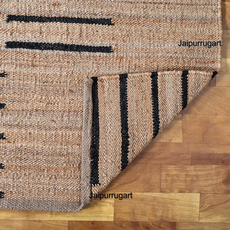 May include: A natural fiber rug with black stripes. The rug is folded in half, showing the underside of the rug. The rug is made of jute and has a natural, earthy color. The rug is perfect for adding a touch of natural beauty to any room.
