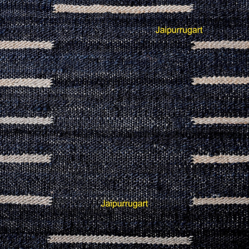 May include: A dark navy blue woven textile featuring a repeating pattern of horizontal cream-colored stripes. The fabric has a visible coarse texture with a grid-like weave. The yellow text Jaipurrugart is printed twice on the surface of the fabric.