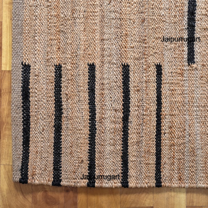 May include: A natural fiber rug with black stripes. The rug is made of jute and has a woven texture. The rug is labeled "Jaipur Rug Art".