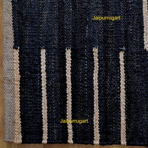 May include: A woven rug featuring a dark navy blue textured surface with vertical stripes in a natural beige tone. The rug has a thick border of the same beige material along the left edge. The text Jaipurrugart is visible in yellow.