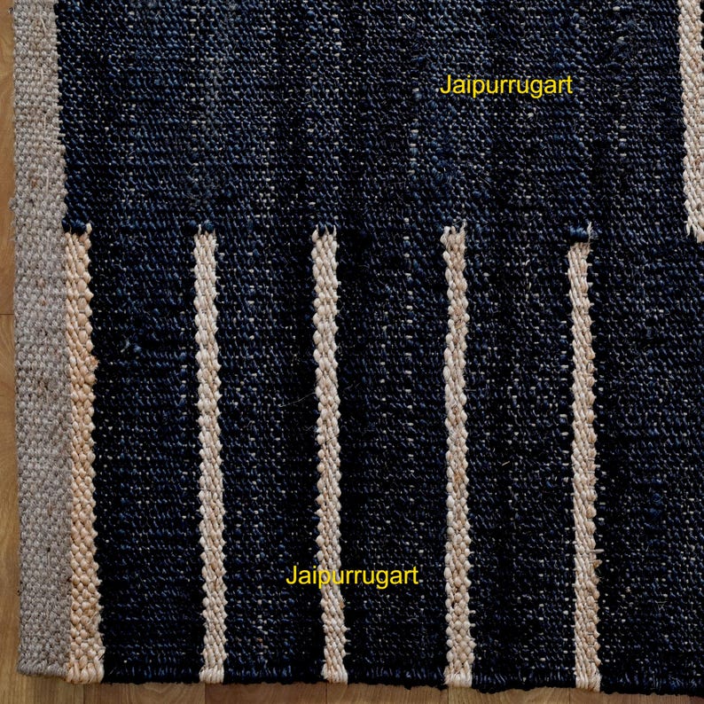 May include: A woven rug featuring a dark navy blue textured surface with vertical stripes in a natural beige tone. The rug has a thick border of the same beige material along the left edge. The text Jaipurrugart is visible in yellow.