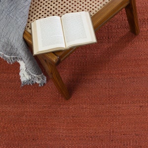 May include: A solid rust-colored woven area rug with a textured surface. A wooden chair with a woven cane seat sits on the rug, holding an open book with white pages and black text. A gray fringed throw blanket is draped over the side of the chair.