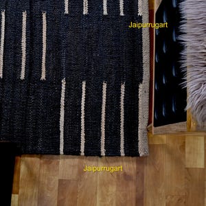 May include: A rectangular woven rug featuring a bold pattern of wide black sections alternating with thin vertical beige stripes. The text Jaipurrugart is visible in yellow on the rug and the wooden floor. The rug has a textured, natural fiber appearance.