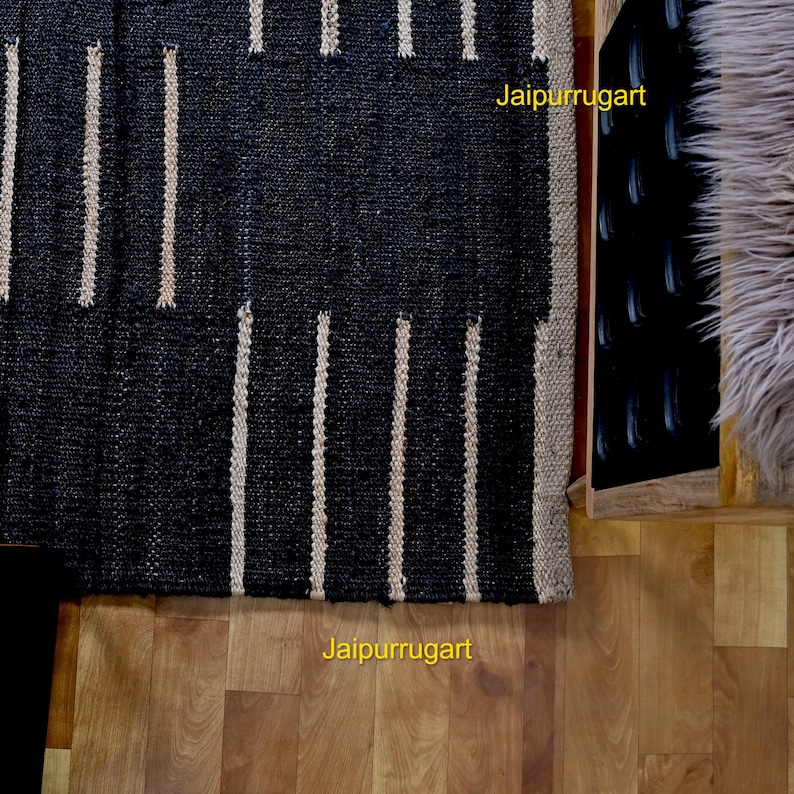 May include: A rectangular woven rug featuring a bold pattern of wide black sections alternating with thin vertical beige stripes. The text Jaipurrugart is visible in yellow on the rug and the wooden floor. The rug has a textured, natural fiber appearance.
