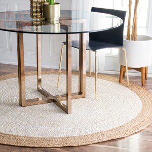 Könnte beinhalten: Ein runder, weißer und naturfarbener Jute-Teppich mit geflochtenem Rand. Der Teppich liegt unter einem Glastisch mit goldfarbenem Metallgestell und einem schwarzen Stuhl.