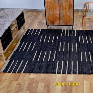 May include: A rectangular area rug featuring a dark black base with a repeating pattern of short, vertical, light beige stripes arranged in staggered columns. The rug is positioned on a light-colored wooden floor, with the text Jaipurrugart visible.