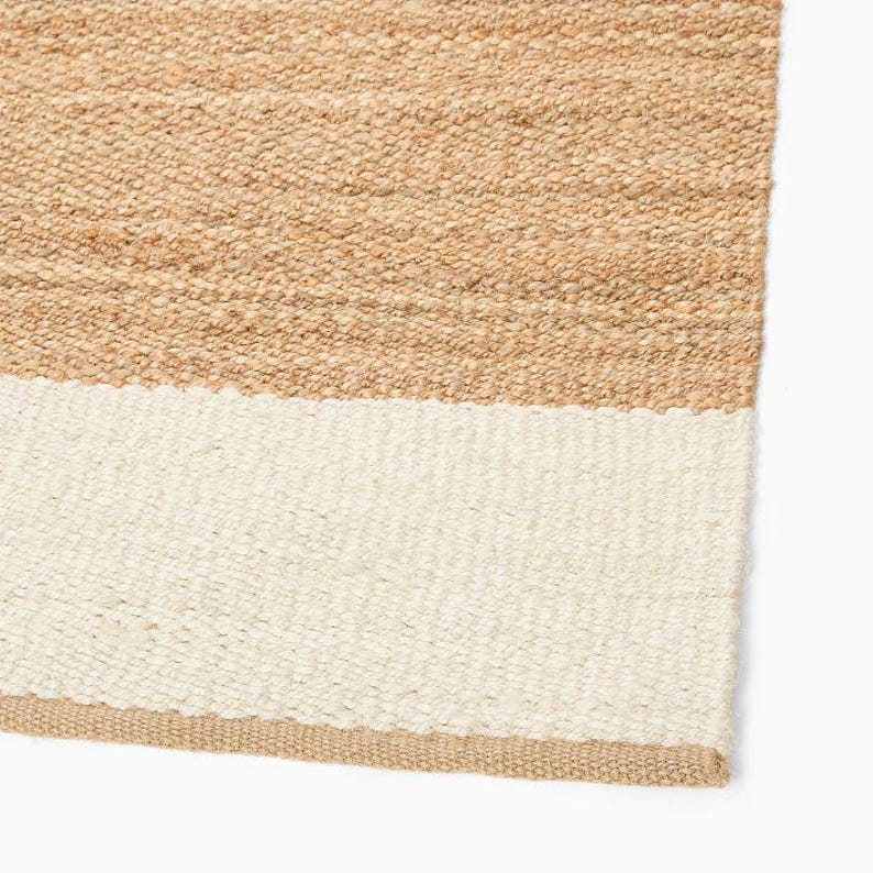 May include: A woven area rug featuring a textured, natural tan jute fiber construction with a wide, solid off-white horizontal stripe running across the lower section. The material displays a dense, braided weave pattern throughout the surface.