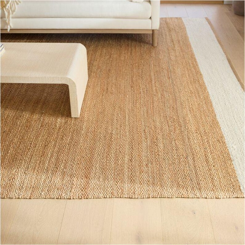May include: A rectangular area rug featuring a woven natural tan jute texture with a wide, solid off-white border along one side. The rug is constructed from thick, braided fibers, creating a consistent, dense, and tactile surface pattern.