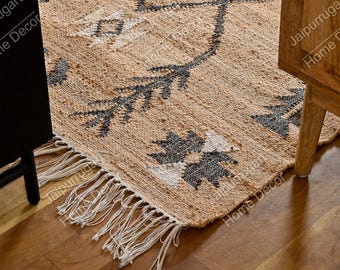 May include: A woven area rug featuring a tan base with dark gray and white geometric patterns. The rug has a textured surface and is finished with long, white fringe tassels along the edge, creating a distinct border detail on the floor.