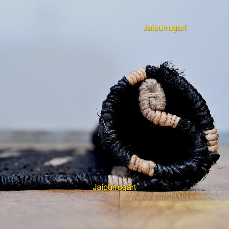 May include: A tightly rolled rug featuring a thick, braided texture in deep black and natural beige fibers. The rug is partially unrolled, revealing a repeating pattern of black and beige woven elements. The text Jaipurrugart is visible.